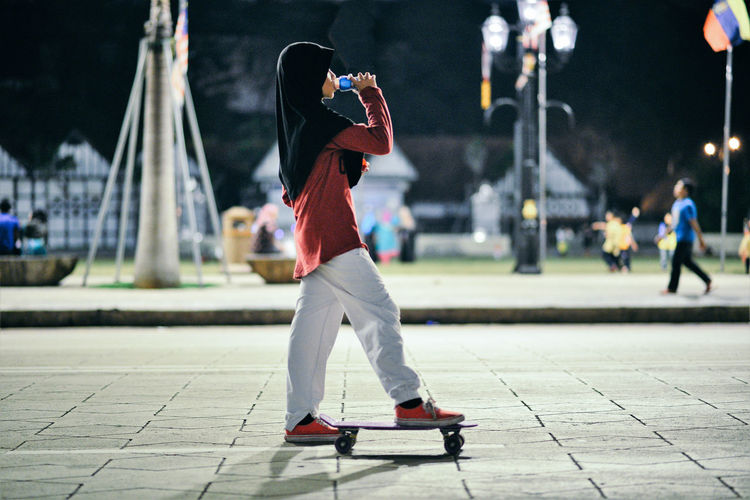 Side view of teenage girl drinking while standing with skateboard on footpath in park at night