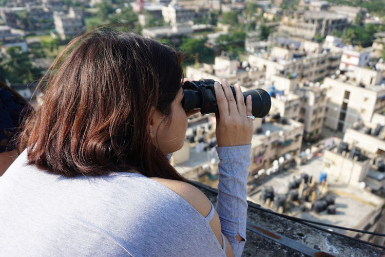 Rear view of woman looking through binoculars against cityscape