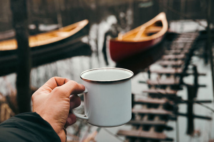 A tin hiking cup in hand with canoe boats in the background, point of view shot. 