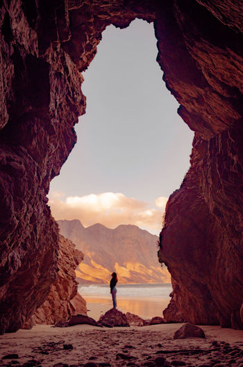 Woman standing under rock formation at beach against sky