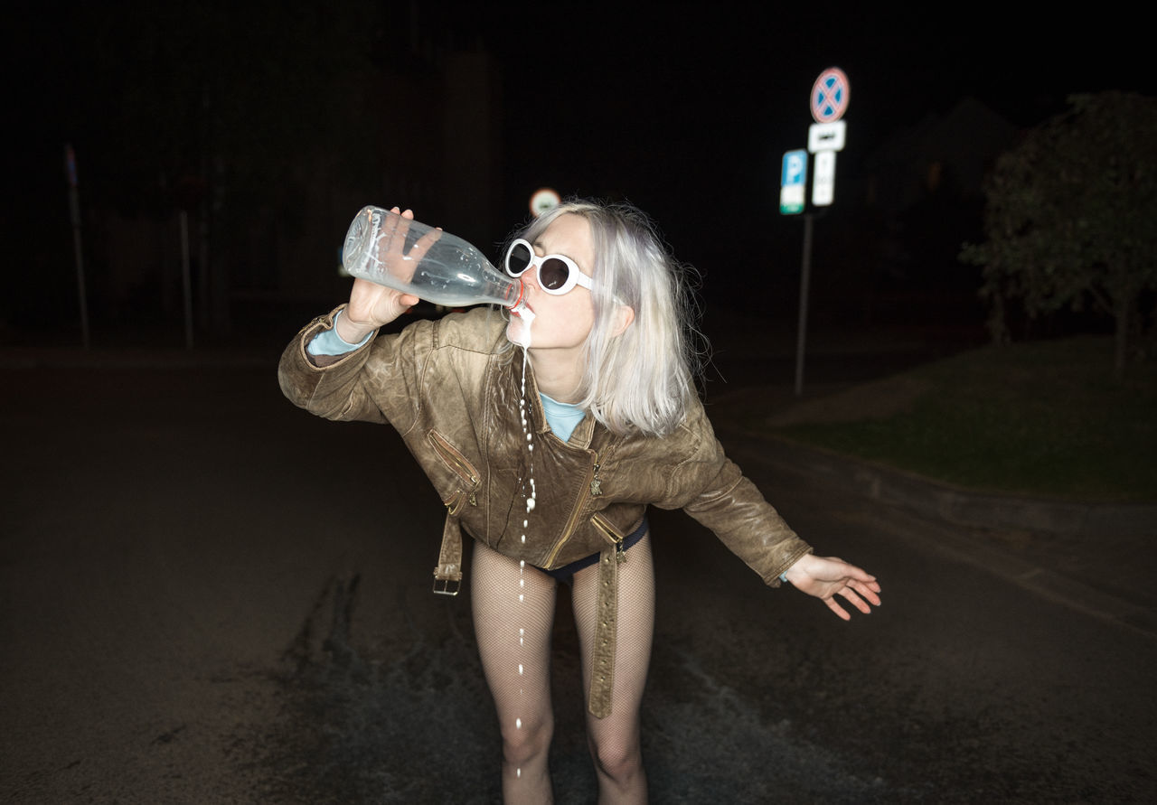 Young woman drinking water while standing on road at night