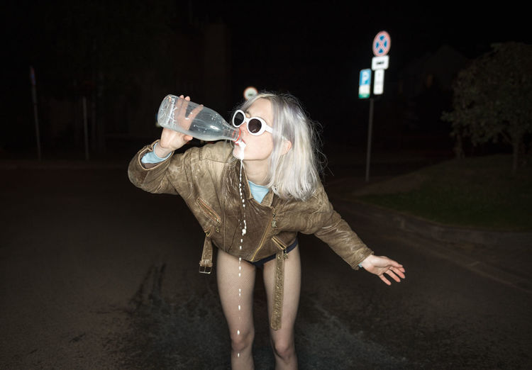 Young woman drinking water while standing on road at night