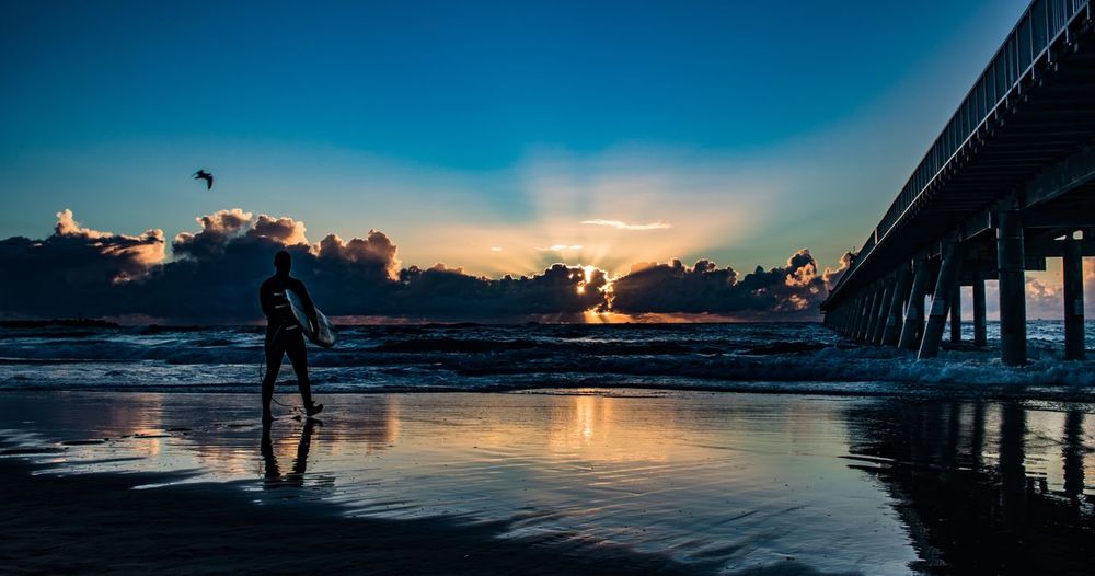 Silhouette man carrying surfboard while walking towards sea on shore at beach against sky during sunset