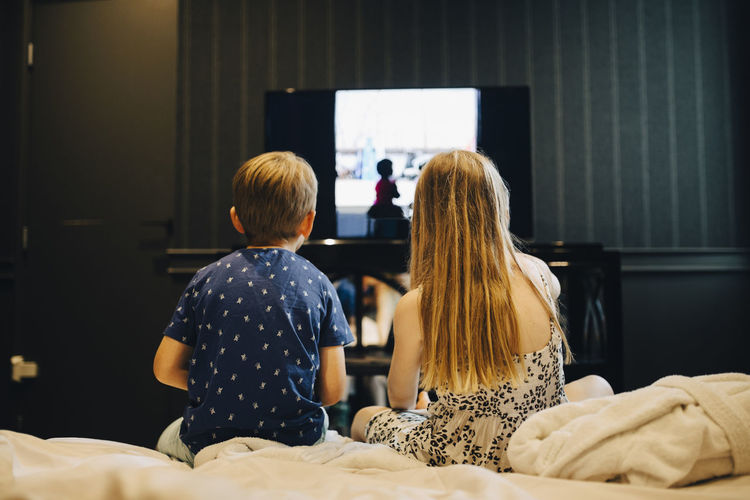 Rear view of siblings watching television while sitting on bed in hotel room