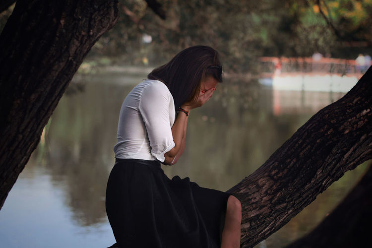 Side view of young woman covering face while sitting on tree trunk