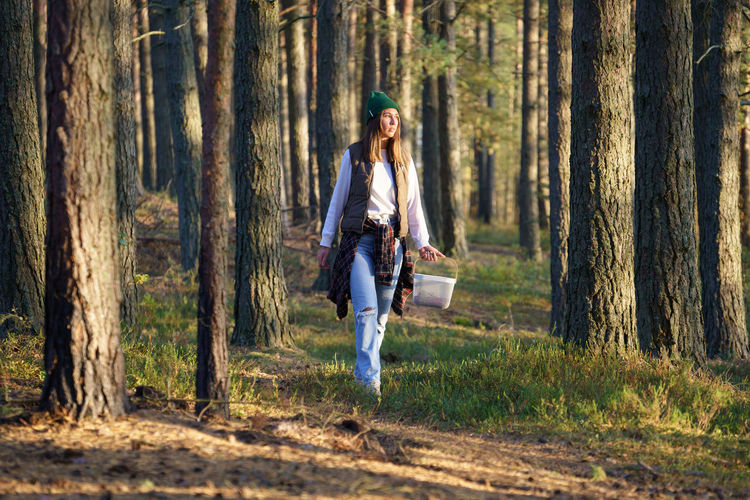 Young urban woman walks in autumn forest among trees searching mushrooms at sunset