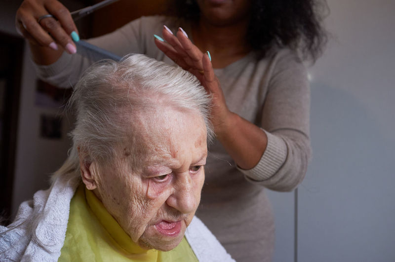 Caring african american woman caregiver, cutting her elderly woman hair at home