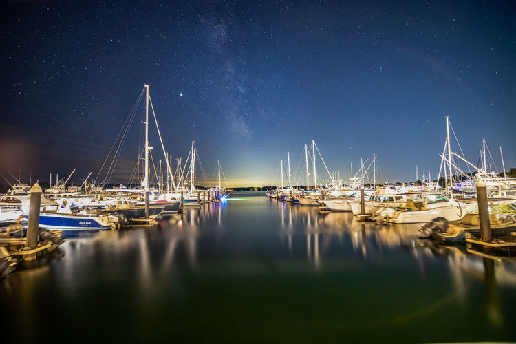 Sail boats docked in the harbor reflecting under the milky way sky.