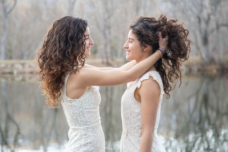 Side view of smiling young woman holding sister hair while standing by lake