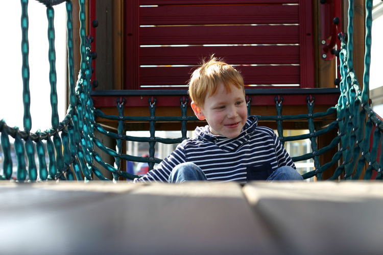 Cute smiling boy looking away while sitting on outdoor play equipment at playground