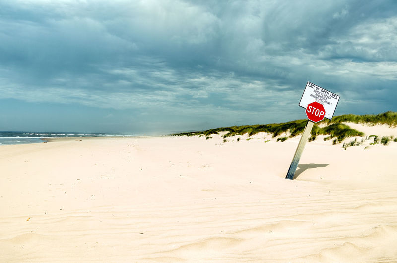 Stop sign on sand at beach against cloudy sky