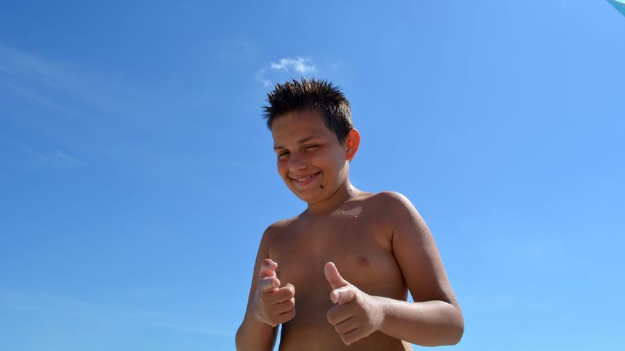 Portrait of shirtless smiling boy gesturing against blue sky