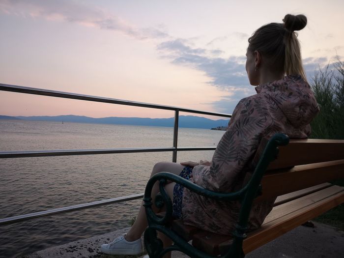 Side view of young woman looking at sea while sitting on bench against sky during sunset