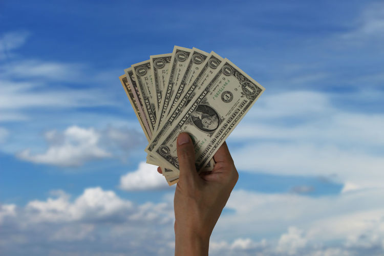 Cropped hand of holding paper currencies against cloudy sky