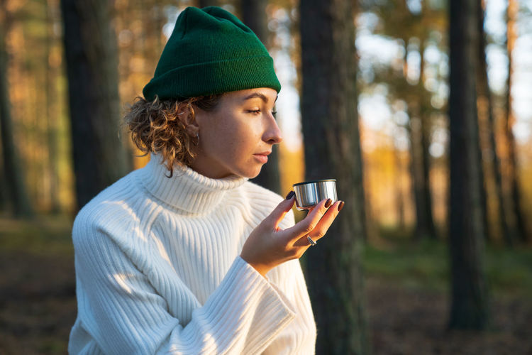 Young woman drink hot tea or coffee from thermos cup walk in autumn forest enjoy nature outside city