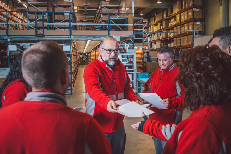 Bearded senior male manager giving instructions on documents to group of employees in red uniform i modern warehouse