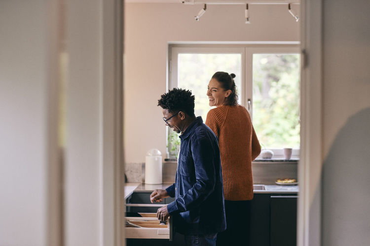 Happy multiracial couple helping each other while standing in kitchen at home