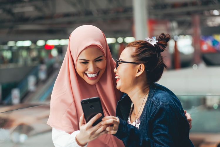 Happy young woman wearing hijab using phone with friend at railroad station
