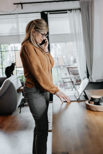 Side view of businesswoman talking on phone while using laptop at home