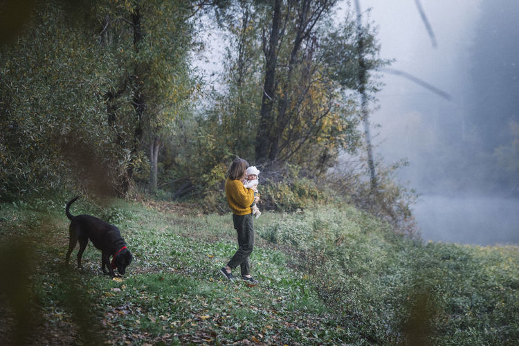 A woman is holding a baby and looking at a dog near a river