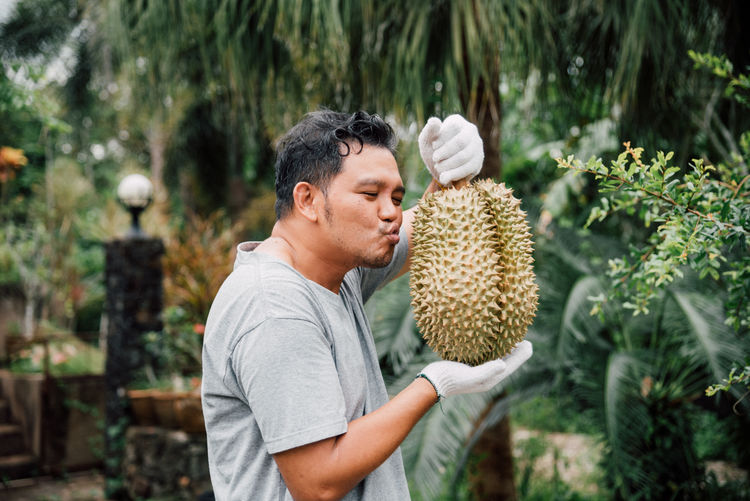 Side view of mature man puckering while holding durian against trees