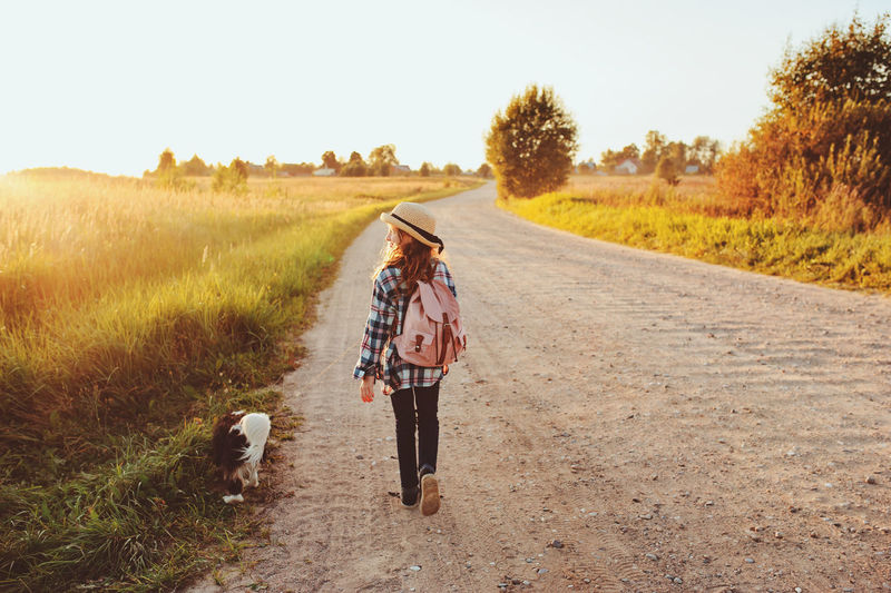 Rear view of girl with dog walking on dirt road against clear sky