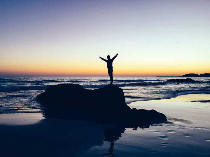 Silhouette person with arms outstretched standing on rock at beach during sunset