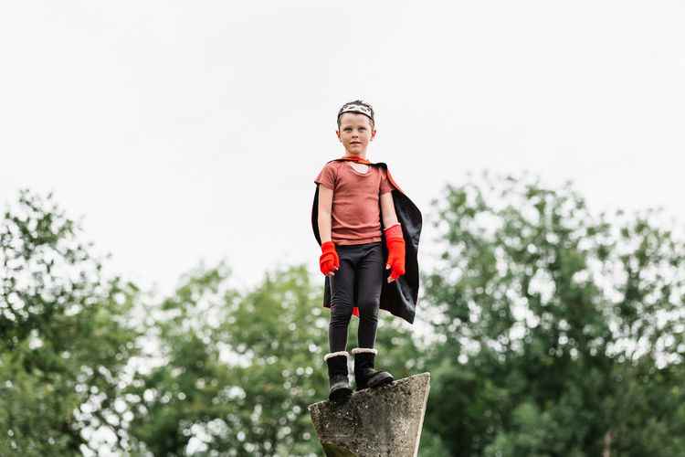 Boy in red superhero cape and hedgehog mask looking away while standing on stone block on blurred background of park trees