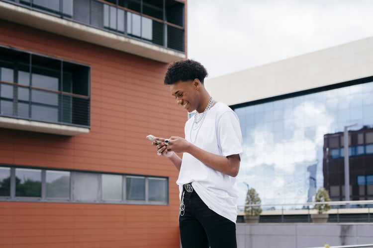 From below side view of happy young african american male student browsing smartphone while walking on city street near modern building
