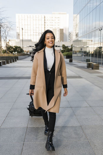 Smiling businesswoman pulling wheeled luggage on footpath