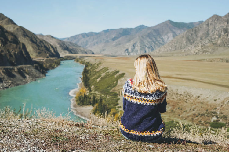 Young blonde woman in nordic sweater sitting on background of turquoise katun river, altai mountains
