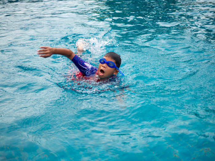 Boy wearing a swimsuit and glasses swimming in the middle of the pool with a blue water background