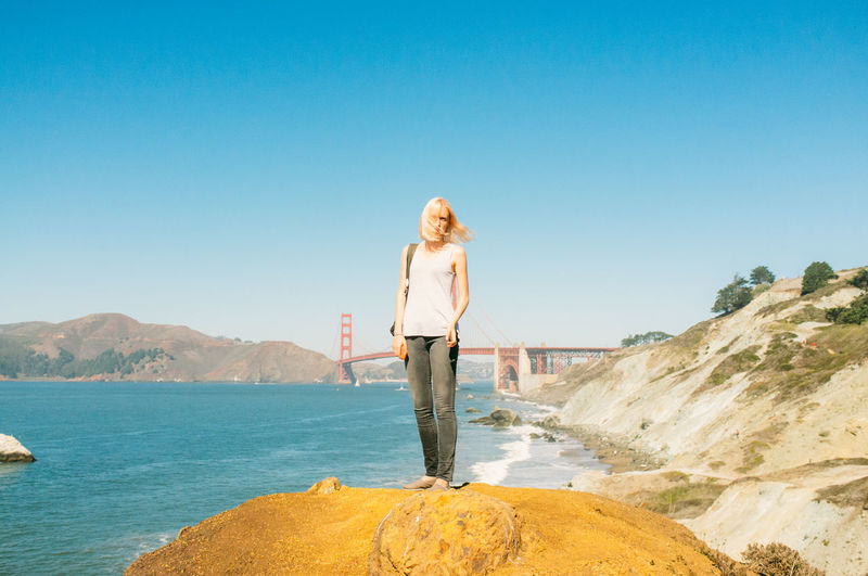 Full length of woman standing on rock at sea shore by golden gate bridge against clear blue sky