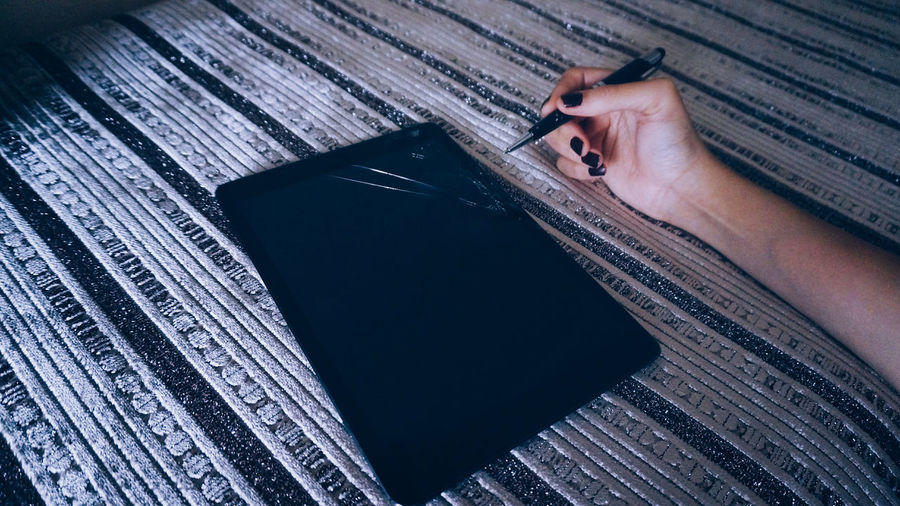 Cropped hand of woman holding pen by broken digital tablet on bed