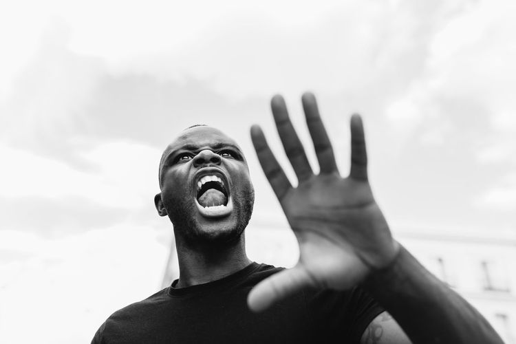 From below black and white of young angry ethnic male screaming and showing stop gesture while looking forward in town