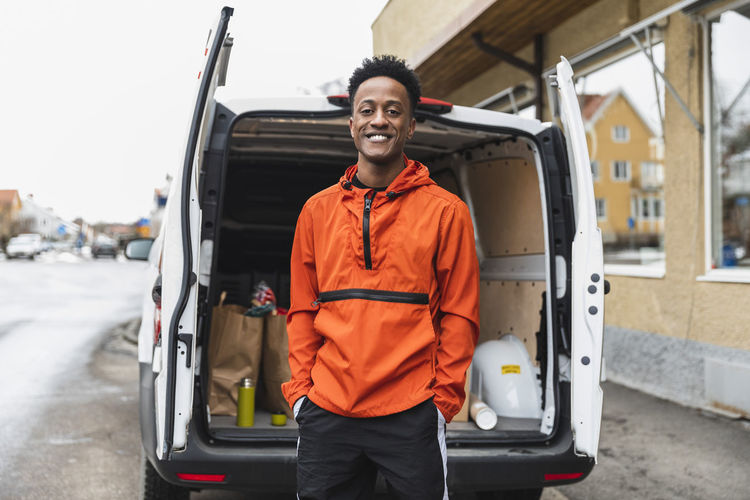 Portrait of smiling essential service man standing with hands in pockets against delivery van