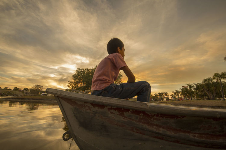 Low angle view of boy sitting in boat moored on lake against cloudy sky during sunset