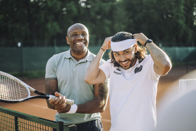 Happy man holding tennis racket with male friend adjusting headband at sports court