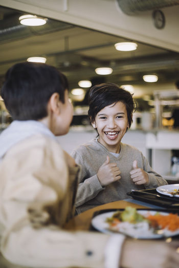 Happy boy showing thumbs up while having food with friend at school cafeteria