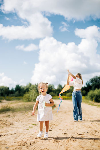 A laughing little girl and her mother run along the road to the field and fly a kite. 