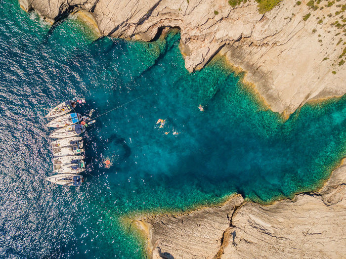 High angle view of boats rafted in remote bay with people swimming in sea and a long jagged coast.