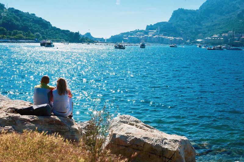 Rear view of people sitting on rock by sea against sky