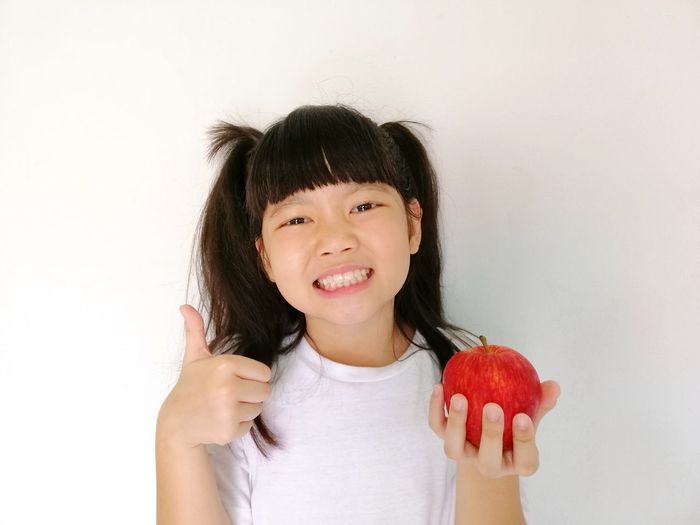 Portrait of smiling girl holding apple while standing against white background