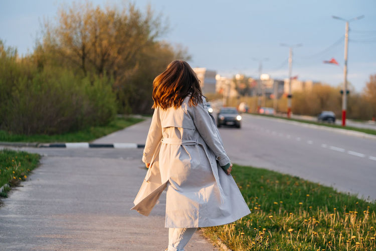 Girl with long hair in a grey trench coat outdoors on the street spring, view from the back