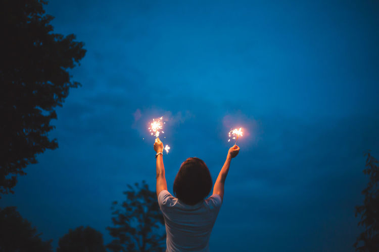Rear view of woman with arms raised holding illuminated sparklers against sky