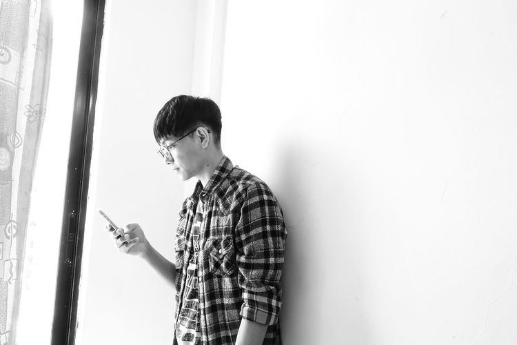 Young man using phone while standing by wall at home