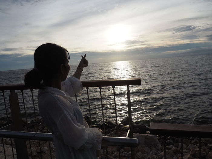 Man sitting on railing by sea against sky during sunset