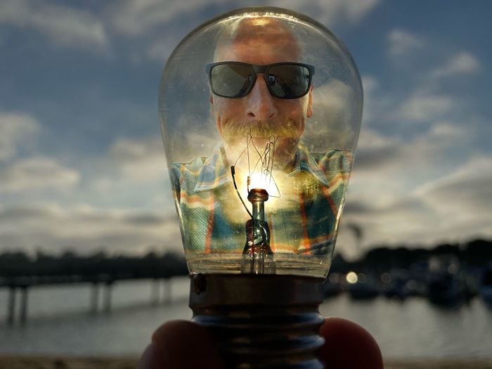 Close-up portrait of man holding light bulb against sky