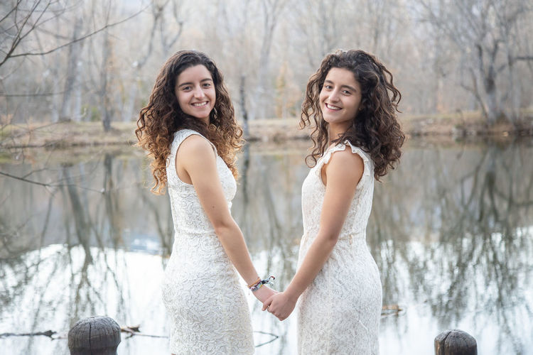Portrait of smiling sisters wearing white dresses while standing by lake