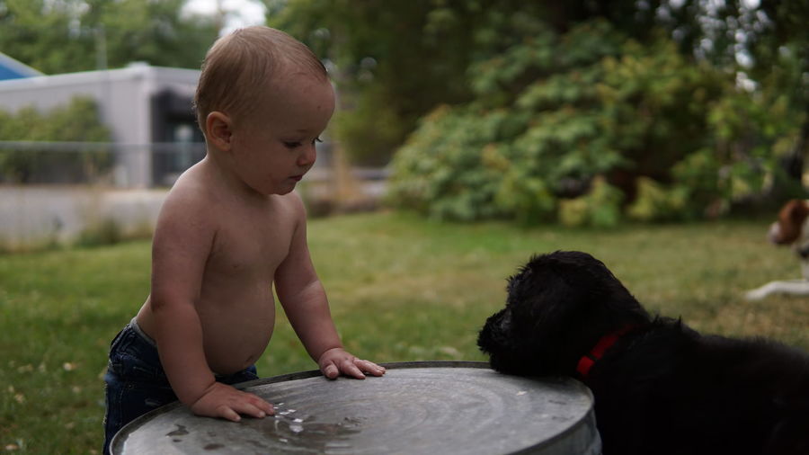 Baby boy in jeans leaning on bucket looking at his puppy.